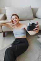 feliz sorrir mulher segurando a velho Câmera e levando As fotos do ela mesma Como uma final de semana fotógrafo dentro casa roupas com uma curto corte de cabelo cabelo sem filtros em uma branco fundo, livre cópia de espaço