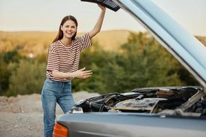 feliz mulher consertado a carro em a rastrear sozinho e sorrisos alegremente perto a aberto de capuz foto