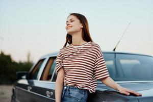 uma jovem mulher dentro uma listrado camiseta e jeans é relaxante a partir de dirigindo, inclinado em dela carro ao lado a estrada e sorridente às a natureza e a pôr do sol foto