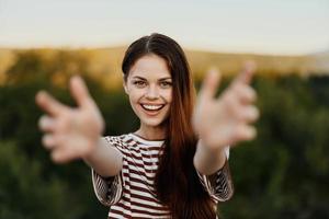 mulher sorridente enquanto olhando às a Câmera e puxar dela mãos para a Câmera fechar-se dentro natureza com uma Visão do a montanhas. feliz viagem estilo de vida Segue mim foto