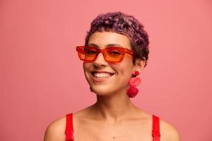 moda retrato do uma mulher com uma curto corte de cabelo dentro colori oculos de sol com incomum acessórios com brincos sorridente em uma Rosa brilhante fundo foto