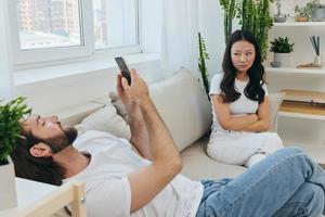 uma homem parece às dele telefone tela e sorrisos durante a argumento com dele namorada. a Bravo e doeu mulher parece dentro dele direção e é triste. família discórdia às lar, telefone vício foto