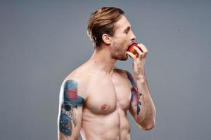 sexy atleta com bombeado acima músculos come a maçã em uma cinzento fundo e uma tatuagem foto