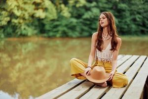 uma jovem hippie mulher senta em uma lago ponte vestindo à moda eco roupas e sorridente foto