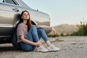uma jovem mulher senta em a terra perto dela carro em a lado do a estrada e parece às a pôr do sol. acampamento depois de uma Difícil estrada viagem foto