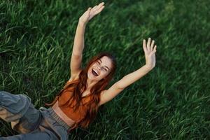 uma mulher deitado dentro natureza em a verde Relva dentro a parque em repouso com dela mãos acima e sorridente com dela boca abrir, dela vermelho cabelo vôo dentro a vento dentro a aberto ar dentro a luz solar. freelancer conceito foto