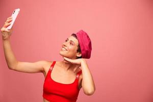 uma jovem mulher blogueiro com colori Rosa cabelo e uma curto corte de cabelo leva uma cenário do ela mesma em a telefone e transmissões uma sorrir dentro à moda roupas e uma chapéu em uma Rosa fundo monocromático estilo foto
