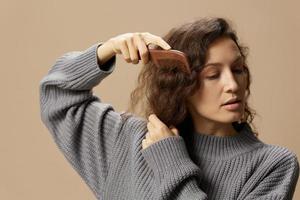 relaxante bonita encaracolado lindo fêmea dentro cinzento casual suéter com escova de cabelo pentes cabelo parece a parte, de lado posando isolado em sobre bege pastel fundo. problemático indisciplinado estragado cabelo conceito. cópia de espaço foto