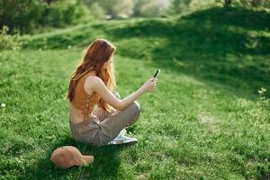 uma jovem feliz mulher sentado dentro a parque em a verde Relva dentro natureza com dela telefone dentro dela mãos dentro dela casual roupas e conversando através da vídeo link, a conceito do saudável lazer meditação foto