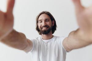 feliz adulto homem com uma barba sorrisos e puxa a cozinhar para dentro a Câmera ouvindo para música dentro fones de ouvido dentro uma angustiado camiseta em uma branco isolado fundo foto