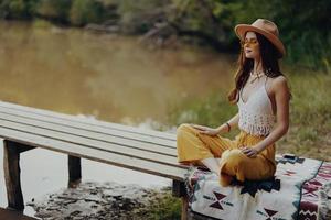 mulher dentro hippie chapéu sorridente e fazendo ioga meditando dentro a lótus posição dentro a pôr do sol luz lado de fora de a lago foto