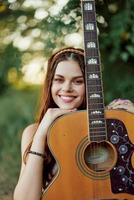 jovem hippie mulher com eco imagem sorridente e olhando para dentro a Câmera com guitarra dentro mão dentro natureza em uma viagem foto