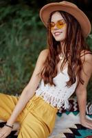 uma jovem hippie mulher dentro roupas ecológicas tem Diversão rindo e encolhendo-se sentado em uma xadrez lado de fora dentro a outono com uma chapéu e amarelo oculos escuros. viagem estilo de vida foto