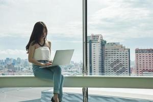 mulher usando um laptop em frente a uma grande janela da cidade foto