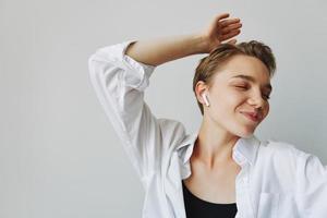 jovem mulher adolescente ouvindo para música com infértil fones de ouvido e dançando lar, sorrindo com dentes com uma curto corte de cabelo dentro uma branco camisa em uma branco fundo. menina natural poses com não filtros foto