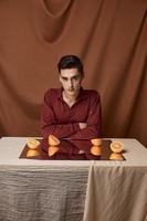 a cara em uma pano fundo senta às uma mesa com uma espelho e maduro laranjas quarto foto