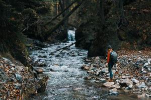 mulher dentro a montanhas perto a rio com uma mochila em dela costas e uma floresta dentro a fundo foto