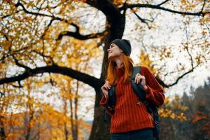 lindo mulher dentro uma vermelho suéter com uma mochila em dela costas perto uma alta árvore dentro outono caído folhas foto
