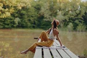 uma hippie mulher senta em uma ponte em a banco do uma rio e goza a lindo cenário por aí dela, feliz dentro a pôr do sol outono Sol. ecológico estilo de vida foto