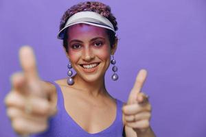 retrato do uma desportivo moda mulher posando sorridente com dentes e apontando uma dedo às a Câmera dentro uma roxa ioga fato de treino e uma transparente boné em uma roxa monocromático fundo foto