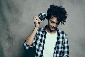 jovem homem com uma Câmera dentro mão e dentro uma xadrez camiseta em uma cinzento fundo dentro de casa fotógrafo foto