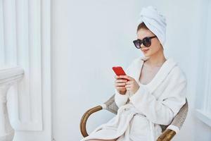 uma mulher dentro uma branco roupão senta dentro uma cadeira com uma vermelho telefone dentro Sombrio óculos aqui ou dentro a manhã foto
