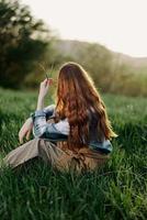 uma mulher senta em a verde Relva dentro uma parque com dela costas para a Câmera e relaxa dentro natureza dentro a verão pôr do sol tarde luz foto