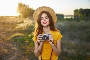 mulher com fechadas olhos com uma Câmera dentro dela mãos vestindo uma chapéu vermelho lábios natureza Sol estilo de vida foto