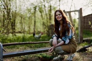 uma jovem mulher agricultor senta às uma pássaro alimentador e Verificações a composição do a grão para alimentação a galinhas dentro uma caneta dentro a campo foto