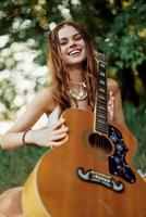 uma jovem hippie mulher com uma guitarra dentro dela mãos sorrisos docemente para dentro a Câmera em uma viagem para natureza estilo de vida dentro harmonia foto