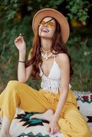 hippie mulher dentro eco roupas risos e mexe Como ela senta em uma xadrez lado de fora dentro a outono com uma chapéu e amarelo oculos de sol foto