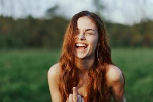 uma jovem mulher sorrisos e parece para dentro a Câmera com dela longo, vermelho, ondulado, brilhante cabelo dentro uma parque com verde Relva dentro a verão pôr do sol. a conceito do saudável vivendo, beleza e cabelo Cuidado. foto