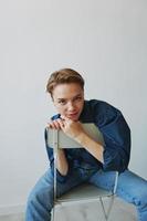 uma jovem mulher sentado dentro uma cadeira às casa sorridente com dentes com uma curto corte de cabelo dentro jeans e uma jeans camisa em uma branco fundo. menina natural poses com não filtros foto