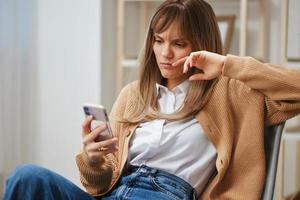 confuso infeliz jovem Loiras senhora dentro caloroso suéter ler mau notícia usar telefone sentado dentro poltrona às moderno casa interior. pausa a partir de trabalhar, levar uma quebrar, social meios de comunicação dentro livre Tempo conceito. cópia de espaço foto