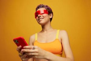 mulher blogueiro sorridente dentro incomum vermelho milenar óculos segurando dela telefone e olhando às a tela contra a laranja fundo dentro uma à moda amarelo topo foto