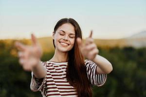 mulher sorridente enquanto olhando às a Câmera e puxar dela mãos para a Câmera fechar-se dentro natureza com uma Visão do a montanhas. feliz viagem estilo de vida Segue mim foto