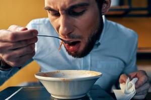 uma bonito homem com uma barba come borscht dentro uma prato às uma mesa dentro uma cafeteria e uma Assistir em dele mão foto