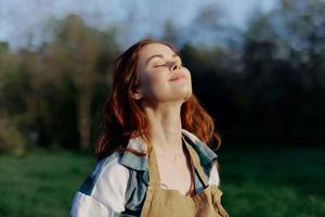 retrato do uma lindo jovem mulher com vermelho cabelo dentro natureza dentro uma parque dentro a verão, olhando às a pôr do sol contra uma fundo do verde Relva e árvores foto