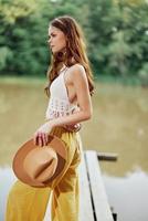 uma jovem mulher sorridente dentro a imagem do uma hippie e vestido ecológico dançando dentro natureza de a lago vestindo uma chapéu e amarelo calça dentro a verão pôr do sol foto