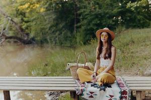 mulher dentro hippie roupas sentado dentro natureza de a lago em uma ponte relaxante e admirando a cenário foto