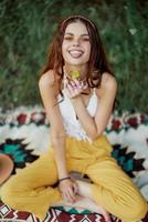 jovem hippie mulher sentado dentro natureza em uma xadrez com uma flor dentro dela mãos e olhando às a Câmera sorridente Atenciosamente dentro roupas ecológicas dentro outono dentro a pôr do sol foto