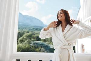 retrato do linda mulher grandes cabelo dentro uma branco roupão de banho ficando em a sacada dentro uma hotel montanha Visão foto