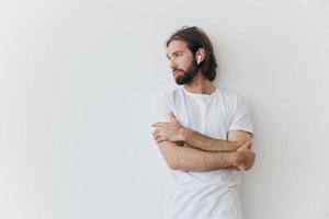 uma homem com uma barba e grandes cabelo dentro uma branco camiseta e azul jeans carrinhos contra uma branco parede, inclinado contra isto e ouvindo para música com sem fio branco fones de ouvido, encarando pensativamente foto