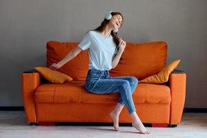 sorridente mulher sentado em a sofá às casa ouvindo para música em fones de ouvido inalterado foto