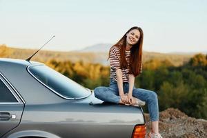 uma mulher carro motorista senta em a tronco do uma carro e sorrisos admirando uma lindo Visão do outono natureza e montanhas foto