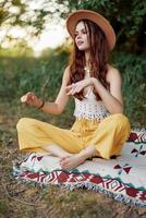 mulher dentro ecológico roupas dentro uma hippie Veja senta em uma colori xadrez meditando dentro a lótus posição sorridente e olhando às a outono pôr do sol dentro a parque foto