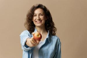 fofa alegre feliz encaracolado lindo fêmea dentro jeans casual camisa puxa maçã às Câmera posando isolado em sobre bege pastel fundo. saudável Comida. natural ecológico produtos conceito. cópia de espaço foto