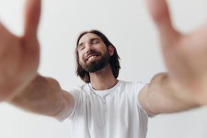 feliz adulto homem com uma barba sorrisos e puxa a cozinhar para dentro a Câmera ouvindo para música dentro fones de ouvido dentro uma angustiado camiseta em uma branco isolado fundo foto