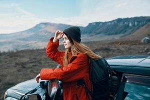 mulher dentro uma chapéu com uma mochila em dela costas inclinou-se em a porta do uma carro dentro a montanhas ao ar livre foto