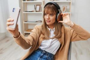 alegre jovem Loiras senhora dentro caloroso suéter dentro fones de ouvido ouço fav canções fazendo selfie vídeo ligar sentado dentro poltrona às moderno casa interior. música Tempo relaxante legal lista de reprodução conceito. cópia de espaço foto